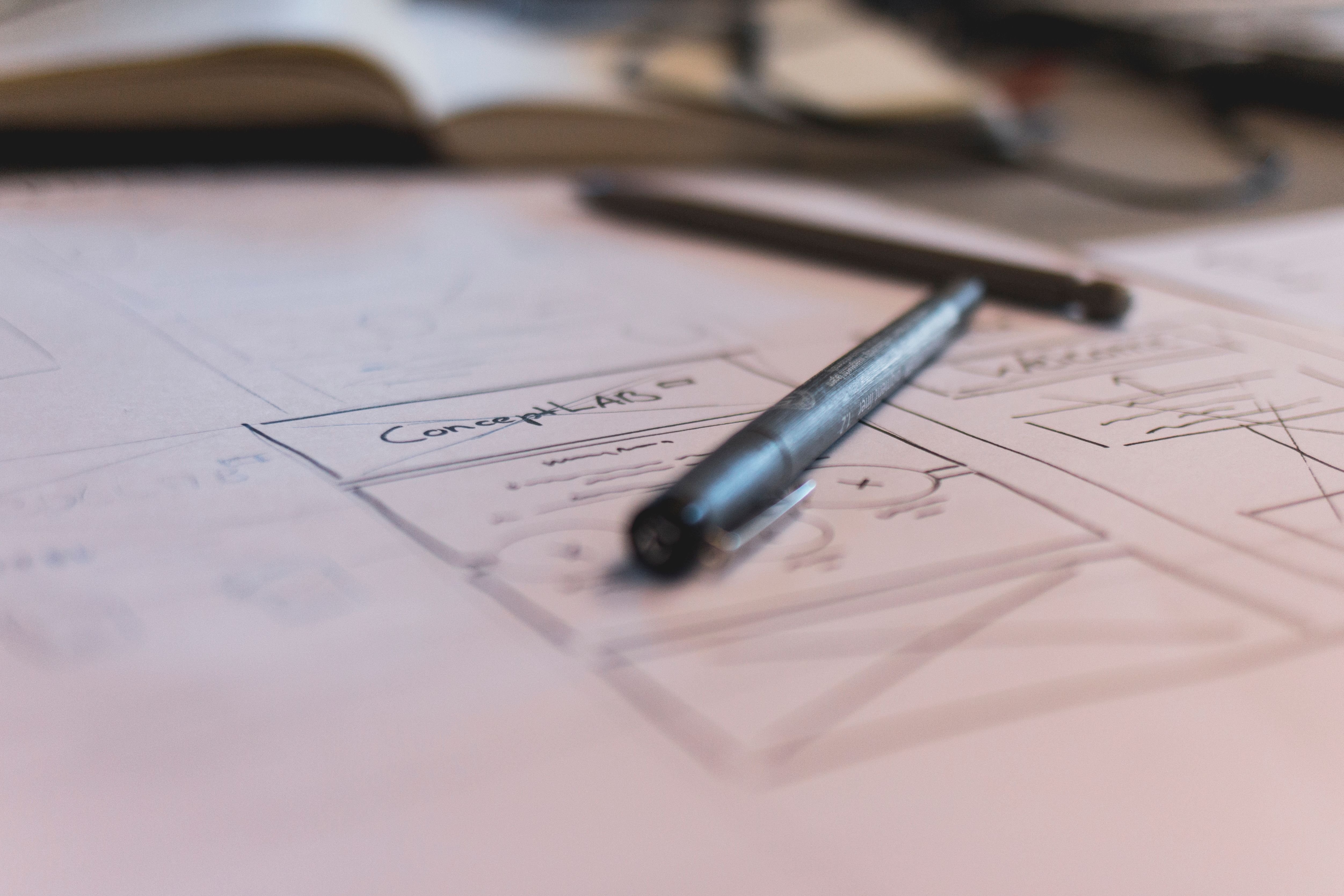 Hand-drawn wireframe sketch with a pen resting on the page, notebook and glasses in the background